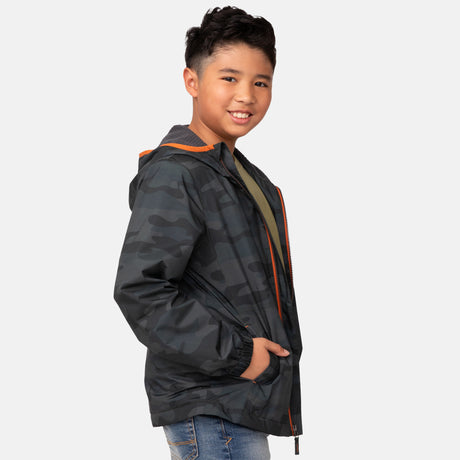 A smiling boy with short dark hair wears the Clearance Boys' Brisk Walk Windshear Jacket in a dark camo print with orange trim and blue jeans, standing with hands in his pockets against a plain light background.