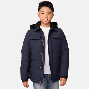 A boy wears a Free Country Boys' Trail Puffer Hoodie Shacket—navy blue and water resistant—paired with a white T-shirt and light blue jeans, standing against a plain white background, facing forward with a neutral expression.