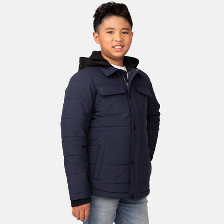 A boy with short dark hair smiles with his hands in the pockets of a navy blue Free Country Boys' Trail Puffer Hoodie Shacket, paired with light blue jeans, standing against a plain white background.