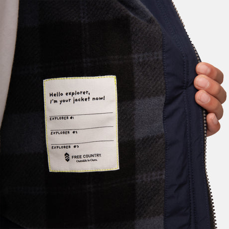 A hand opens a dark plaid Boys' Trail Puffer Hoodie Shacket by Free Country, showing a sewn-in label that says "Hello explorer, I'm your jacket now!" with spaces for Explorer #1-3 and the Free Country logo at the bottom.