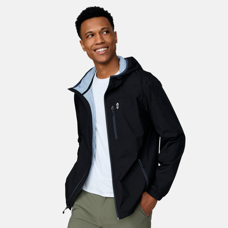 [Josiah is 6'3" wearing a size Medium.] Man wearing the Free Country Men's Agile II Windshear Jacket in Black, size M, made from lightweight wind and water resistant recycled polyester fabric and featuring an attached hood, inner phone pocket and elastic cuffs, styled with a black t shirt and black pants #color_ black