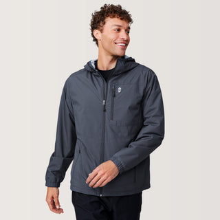 A man with curly hair smiles while wearing the Free Country Men's Agile II Windshear Jacket in dark gray over a black shirt. Featuring zippered chest and side pockets, this lightweight jacket is ideal for everyday wear. He stands against a plain light background.