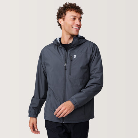 A man with curly hair smiles while wearing the Free Country Men's Agile II Windshear Jacket in dark gray over a black shirt. Featuring zippered chest and side pockets, this lightweight jacket is ideal for everyday wear. He stands against a plain light background.