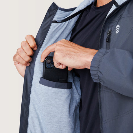 A man in the Free Country Men's Agile II Windshear Jacket, featuring a light gray lining, slips a smartphone into the chest pocket over his black shirt.