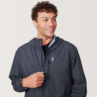 A smiling man with curly hair wears the Free Country Men's Agile II Windshear Jacket in dark gray. He looks to his left while pulling up the zipper with one hand. The background is plain and light-colored.