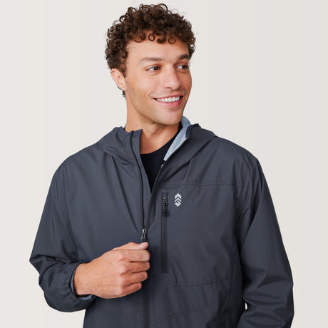 A smiling man with curly hair wears the Free Country Men's Agile II Windshear Jacket in dark gray. He looks to his left while pulling up the zipper with one hand. The background is plain and light-colored.