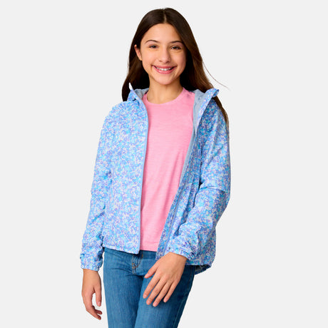A smiling girl with long brown hair wears the Clearance Girls' High Stretch Windshear Jacket in light blue floral over a pink t-shirt and blue jeans, standing against a plain light gray background.