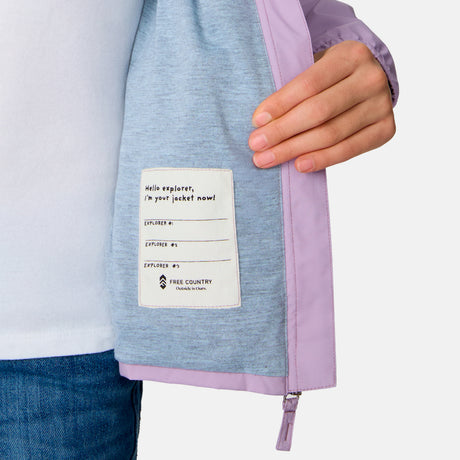 A person holds open a light purple Girls' High Stretch Windshear Jacket by Clearance, revealing an inside label with spaces to write names labeled Explorer #1, Explorer #2, and Explorer #3. The jacket is worn over a white shirt and blue jeans.