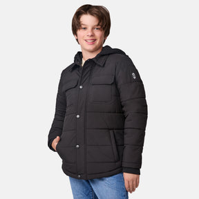 A smiling boy with medium brown hair models the Clearance Boys' Trail Puffer Hoodie Shacket in black—a padded jacket featuring snap buttons and recycled synthetic fill—paired with blue jeans, against a plain white background.