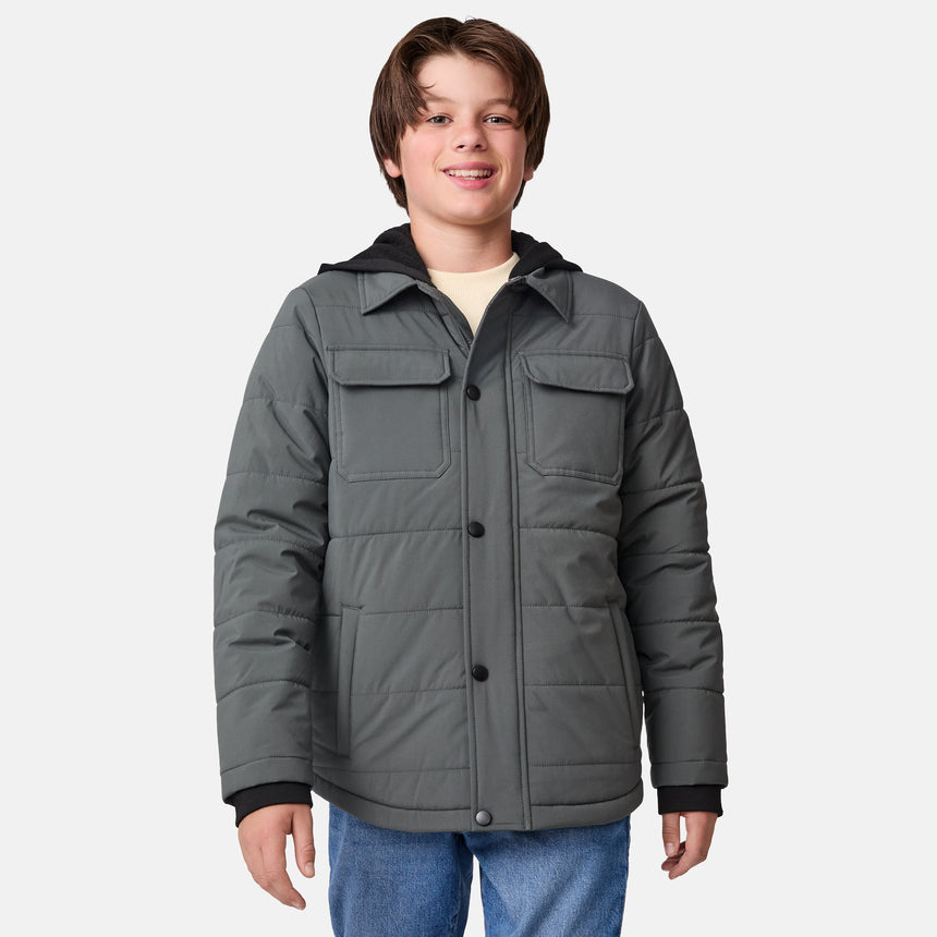 [River is wearing size M.] Model wearing a Free Country dark grey quilted hooded jacket featuring flap chest pockets, side hand pockets, and a partial front zip closure, paired with light innerwear and blue jeans—styled for casual outdoor comfort.