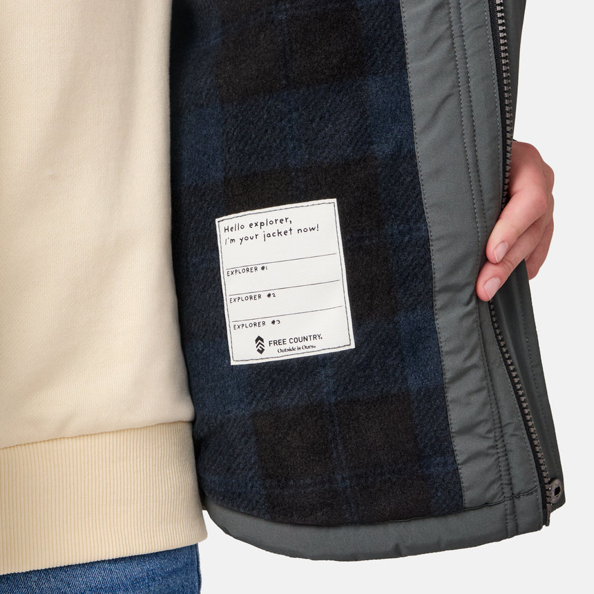 [River is wearing size M.] Model wearing a Free Country dark grey quilted hooded jacket featuring flap chest pockets, side hand pockets, and a partial front zip closure, paired with light innerwear and blue jeans—styled for casual outdoor comfort.