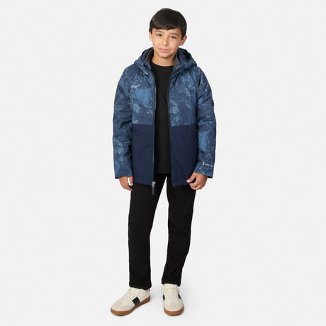 A boy wears a Boys’ High Stretch 3-in-1 Systems Jacket featuring a blue camo-print upper and solid blue lower section over a black shirt and black pants.
