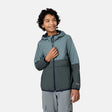 A smiling boy with curly hair stands against a plain white background, zipping up the Free Country Boys' Easy Hiking Colorblock Windshear Jacket in teal and gray, paired with gray pants.