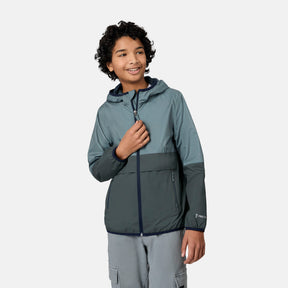 A smiling boy with curly hair stands against a plain white background, zipping up the Free Country Boys' Easy Hiking Colorblock Windshear Jacket in teal and gray, paired with gray pants.