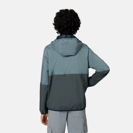 A person with short curly hair, seen from behind, wears the Free Country Boys' Easy Hiking Colorblock Windshear Jacket—blue and gray—and light gray cargo shorts, standing before a plain light background.