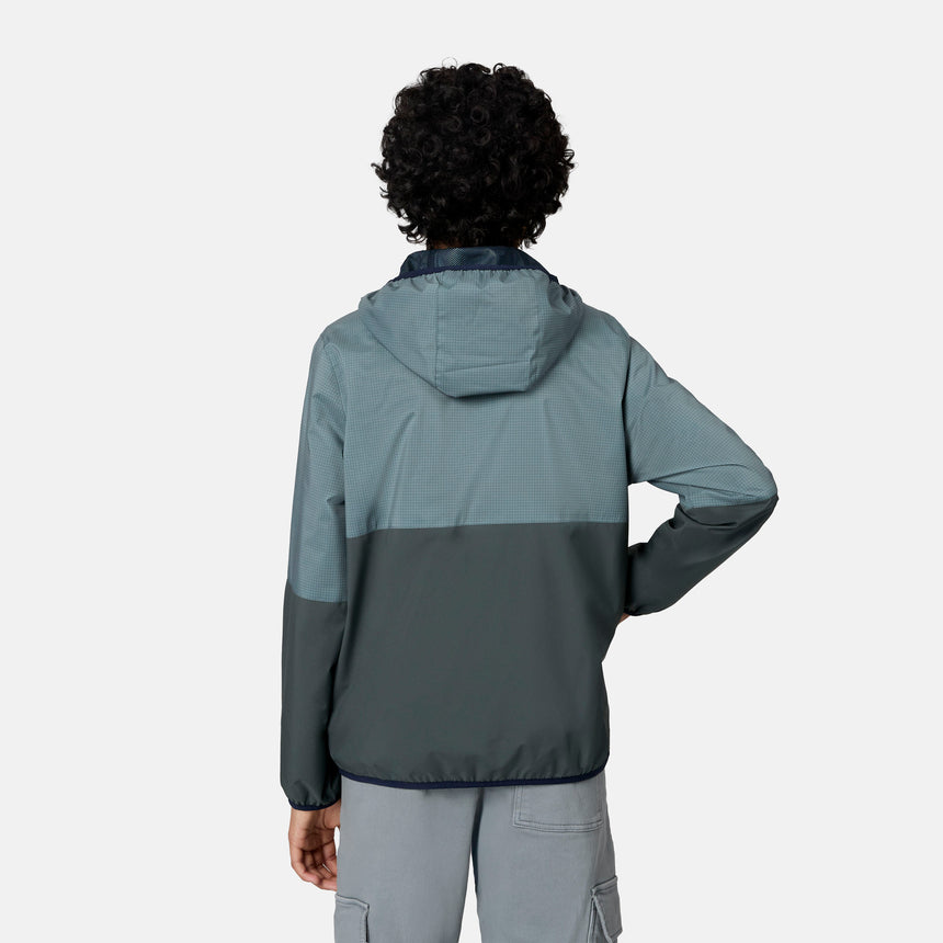 A person with short curly hair, seen from behind, wears the Free Country Boys' Easy Hiking Colorblock Windshear Jacket—blue and gray—and light gray cargo shorts, standing before a plain light background.