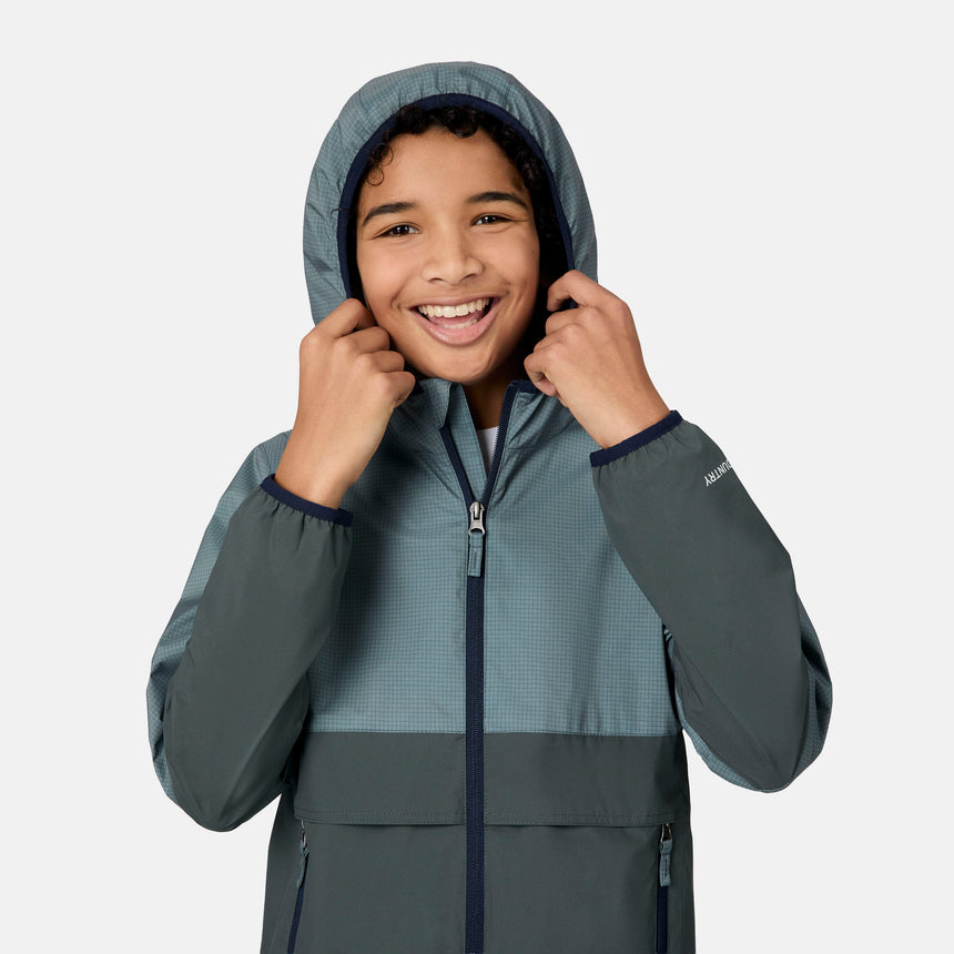 A smiling child with curly hair wears the Free Country Boys' Easy Hiking Colorblock Windshear Jacket, holding the hood with both hands against a plain white background.