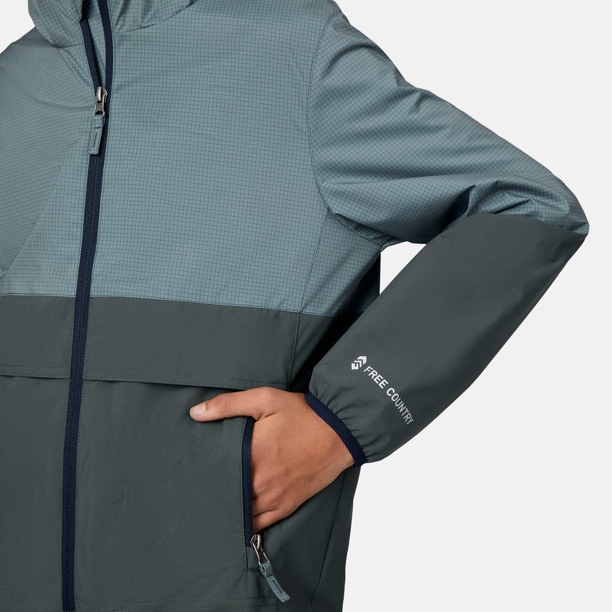 A boy wears the Free Country Boys' Easy Hiking Colorblock Windshear Jacket in two-tone blue, with a front zipper, one hand in the pocket, logo on the sleeve, and a subtle grid pattern on the upper section.