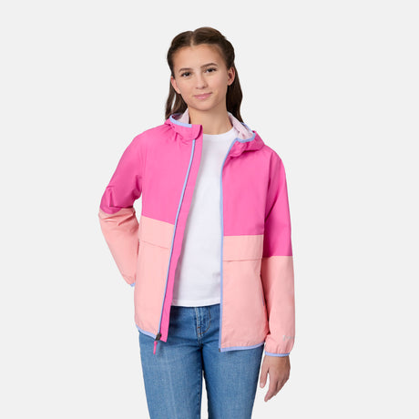 A young person with brown hair stands against a white background, smiling slightly with hands by their sides, wearing the Free Country Girls' Easy Hiking Windshear Jacket in pink over a white shirt and blue jeans.