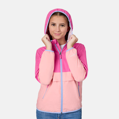 A young woman with light brown hair smiles at the camera, holding the hood of her Free Country Girls' Easy Hiking Windshear Jacket, styled casually with blue jeans against a plain background.