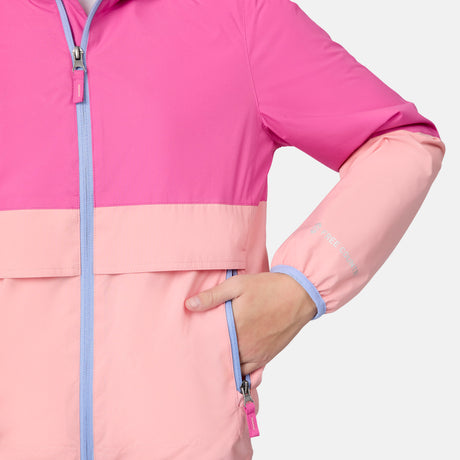 A person wears the Free Country Girls' Easy Hiking Windshear Jacket, a two-tone pink lightweight windbreaker with a light blue zipper, featuring darker pink on top and lighter pink at the bottom, and stands with one hand in the pocket.