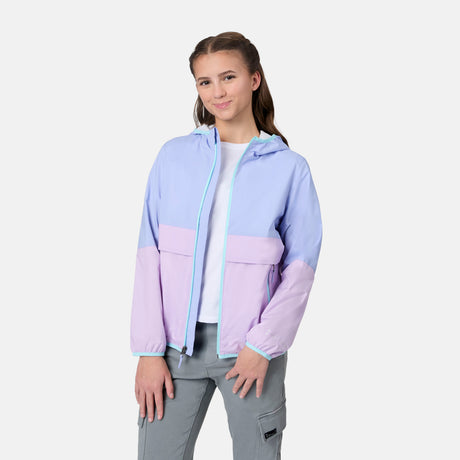 A young woman with brown braided hair wears the Free Country Girls' Easy Hiking Windshear Jacket in light blue and lavender over a white t-shirt and gray cargo pants, standing against a plain background with a slight smile.