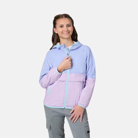 A smiling young woman wears the Free Country Girls' Easy Hiking Windshear Jacket in light blue and lavender with gray cargo pants, standing against a plain white background.