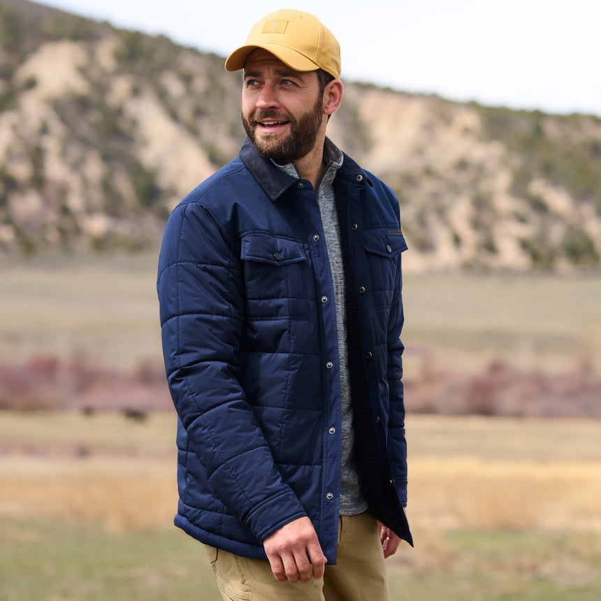 Man wearing a blue puffer jacket and yellow cap standing in a natural landscape.