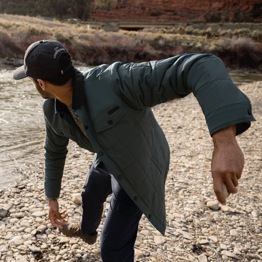 Man in a green coat and black cap standing on a rocky path with a natural landscape in the background