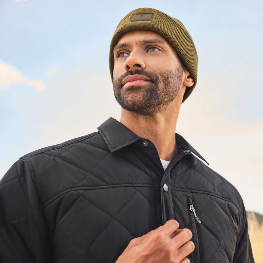 Man wearing a black quilted jacket and green beanie against a blue sky.