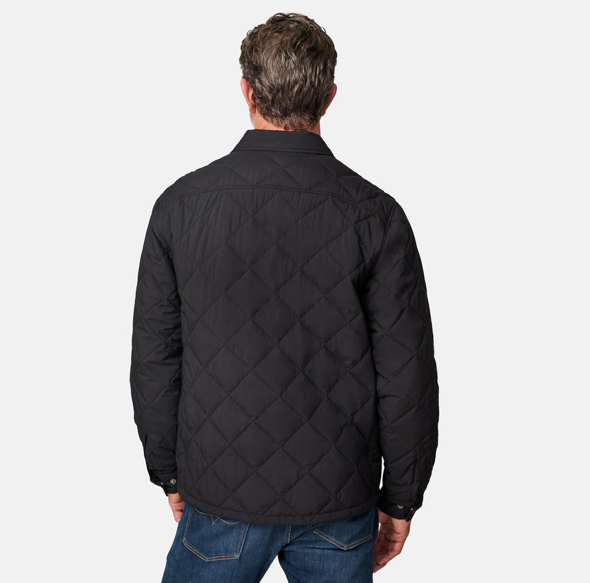 [Kolby is 6'2" wearing size M.] Men’s Free Country black quilted jacket with diamond pattern stitching—featuring button front closure, zippered left chest pocket, and side hand pockets. Layered over a green crewneck tee and blue jeans for a laid-back, outdoors-ready aesthetic.