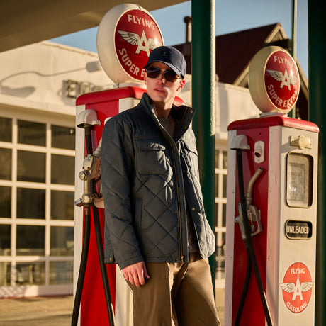 A man wearing a dark gray quilted puffer jacket, brown pants, sunglasses, and a navy cap stands at a vintage red and white Flying A gas station, posing between two retro pumps.