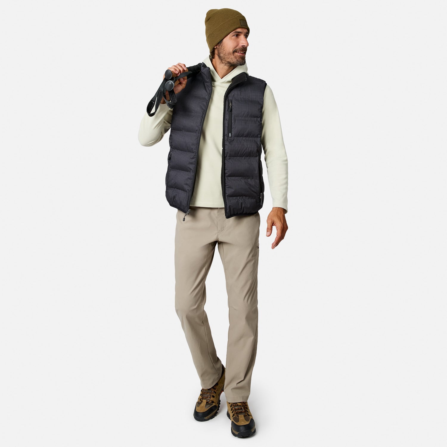 A man wearing a brown beanie, beige long-sleeve shirt, Free Country Men’s Haven Stitchless Reversible Vest for lightweight warmth, tan pants, and hiking boots holds binoculars over his shoulder and looks to the side, smiling.