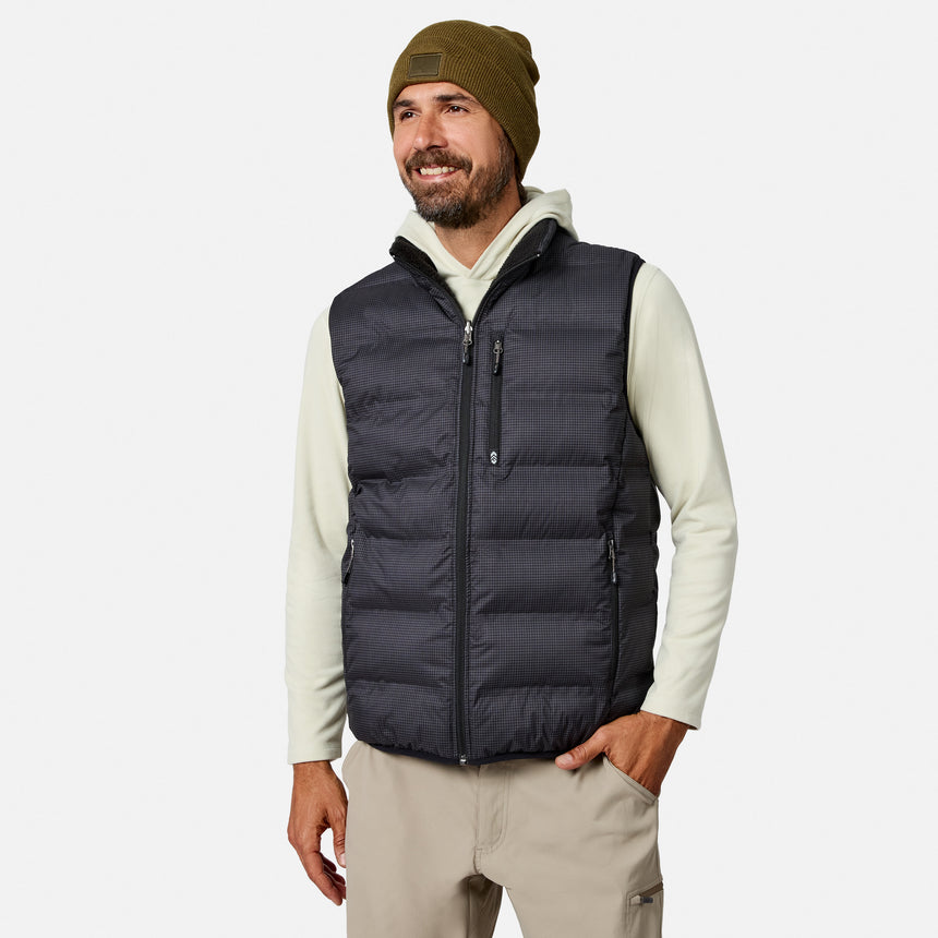 A man stands smiling with one hand in his pocket against a plain white background, wearing the Free Country Men’s Haven Stitchless Reversible Vest in black for lightweight warmth over a light hoodie, paired with tan pants and a green beanie.
