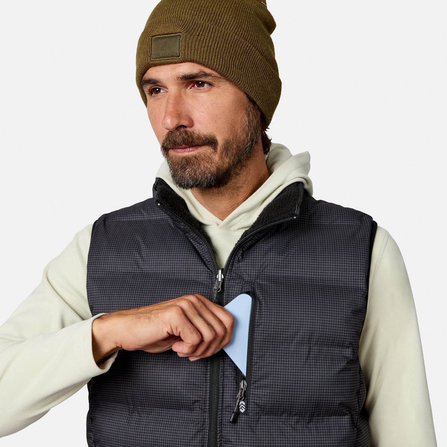 A man in a beanie and the Free Country Men’s Haven Stitchless Reversible Vest, renowned for its lightweight warmth, places a light blue object into the chest pocket of his vest. He looks to the side with a neutral expression against a plain background.