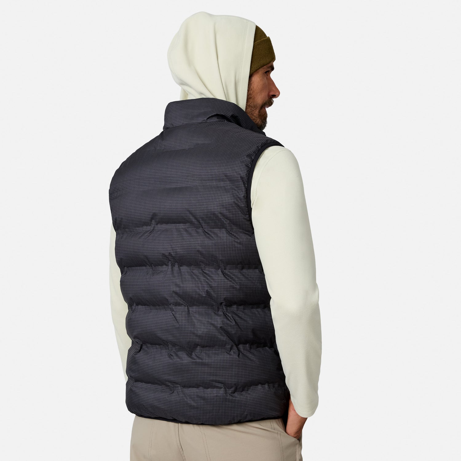 A man facing away wears a cream hoodie, a dark beanie, and the Free Country Men’s Haven Stitchless Reversible Vest—a puffy black vest designed to provide lightweight warmth—against a plain white background.