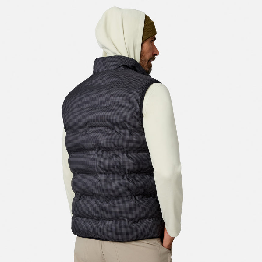 A man facing away wears a cream hoodie, a dark beanie, and the Free Country Men’s Haven Stitchless Reversible Vest—a puffy black vest designed to provide lightweight warmth—against a plain white background.