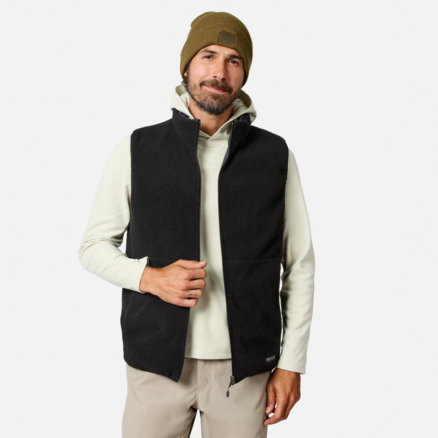 A man wearing a brown beanie, light-colored hoodie, beige pants, and the Free Country Men’s Haven Stitchless Reversible Vest stands against a plain white background, looking at the camera and smiling slightly.
