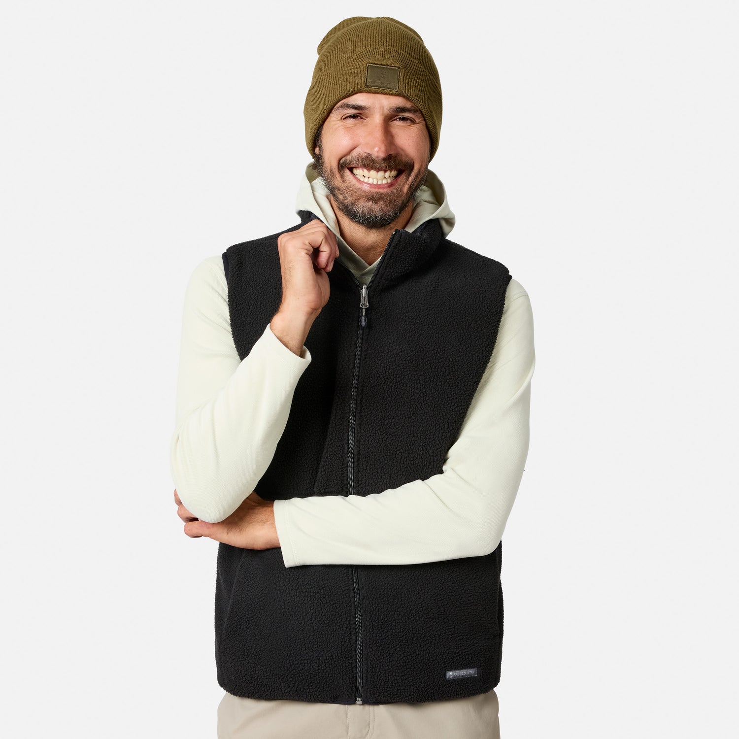 A smiling man stands with arms crossed against a plain white background, wearing a beige knit beanie, light long-sleeve shirt, and the Free Country Men’s Haven Stitchless Reversible Vest—the lightweight vest features a cozy sherpa reverse for added warmth.