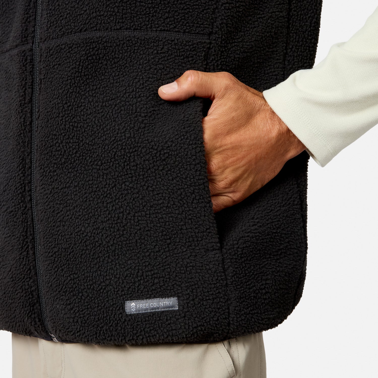A man wears the Free Country Men’s Haven Stitchless Reversible Vest in black, paired with light-colored pants, placing his right hand into one of the vest’s side pockets. This lightweight, warm vest features a small Free Country label near the hem. The background is plain white.