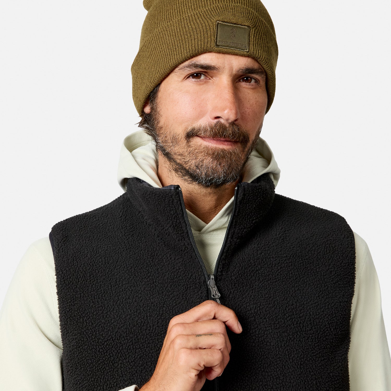 A man with a trimmed beard wears a brown beanie, a light hoodie, and the Free Country Men’s Haven Stitchless Reversible Vest in black. He looks at the camera while holding the vest’s zipper. The vest provides lightweight warmth and features a stylish sherpa reverse. The background is plain and light-colored.