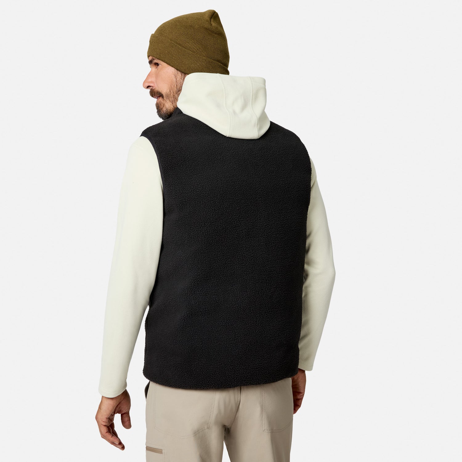 A man stands against a plain white background, facing away, wearing the Free Country Men’s Haven Stitchless Reversible Vest in black with a sherpa reverse for lightweight warmth, layered over a white hoodie, paired with tan pants and an olive green beanie.