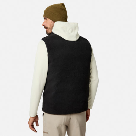 A man stands against a plain white background, facing away, wearing the Free Country Men’s Haven Stitchless Reversible Vest in black with a sherpa reverse for lightweight warmth, layered over a white hoodie, paired with tan pants and an olive green beanie.