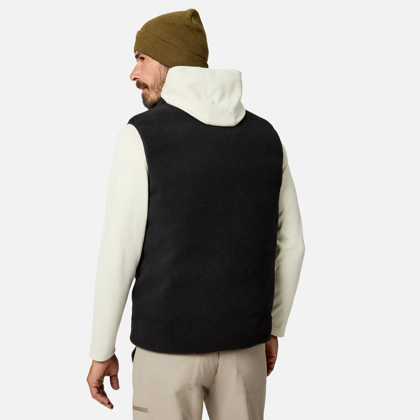 A man stands against a plain white background, facing away, wearing the Free Country Men’s Haven Stitchless Reversible Vest in black with a sherpa reverse for lightweight warmth, layered over a white hoodie, paired with tan pants and an olive green beanie.