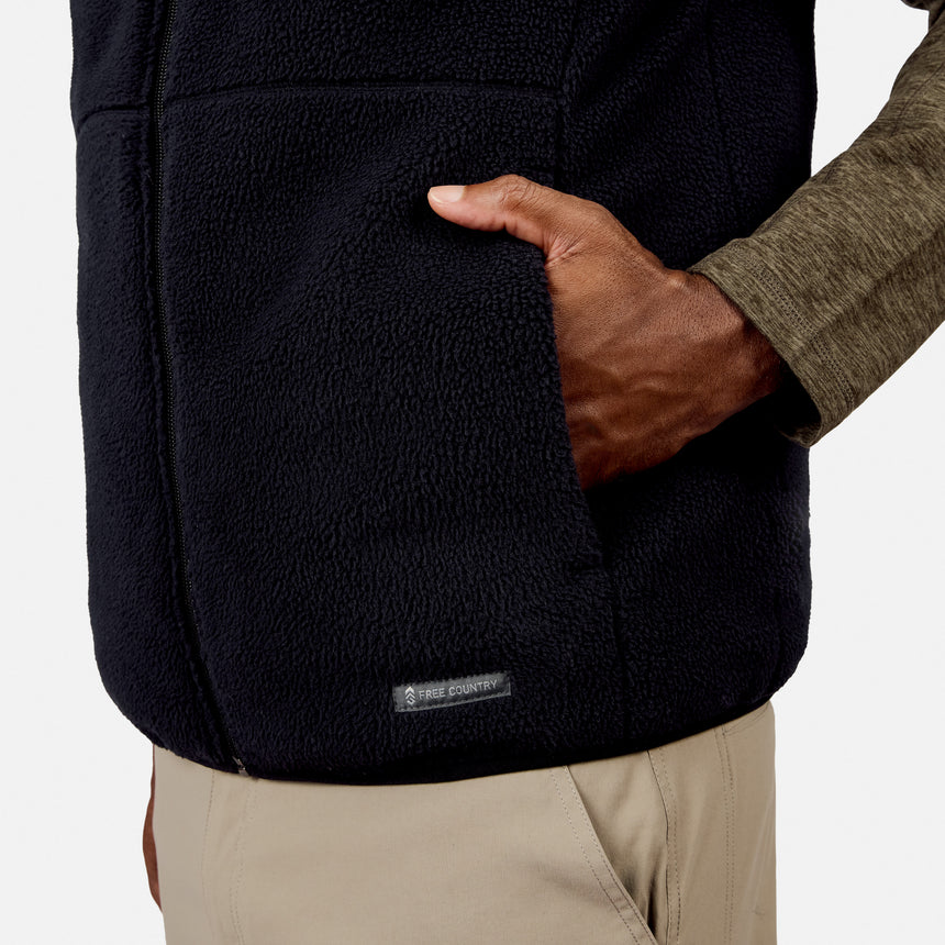A person wearing the Free Country Men’s Haven Stitchless Reversible Vest in black and khaki pants, with one hand in a vest pocket, showing a partial view of the arm and torso.