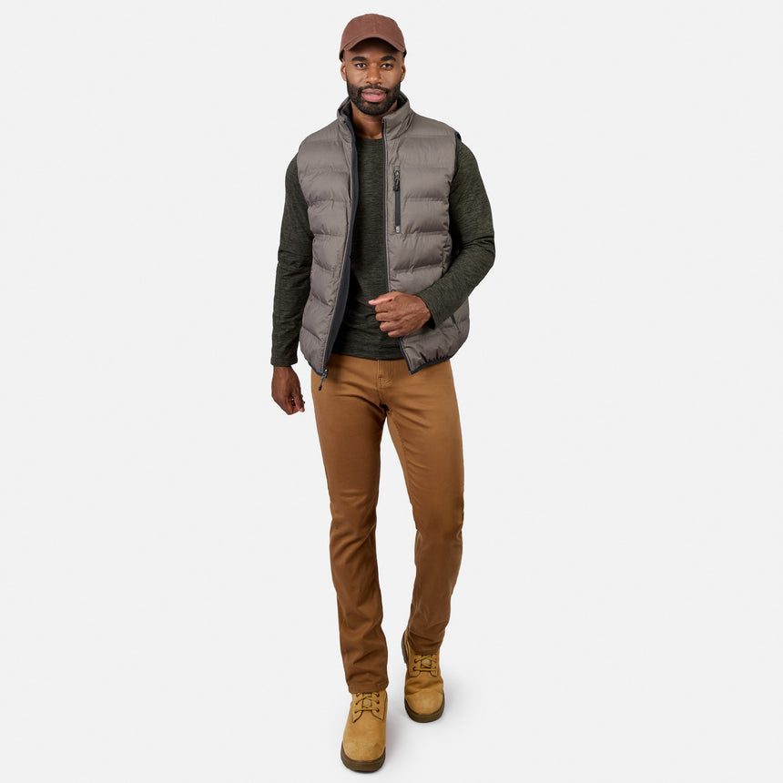 A man wearing a brown cap, the Free Country Men’s Haven Stitchless Reversible Vest for lightweight warmth in gray, a dark green long-sleeve shirt, tan pants, and tan boots stands against a plain white background, looking forward and walking confidently.