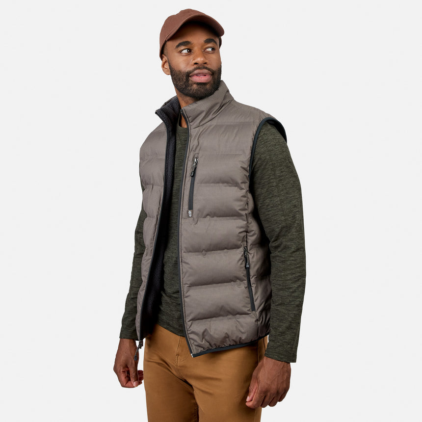 A man wearing a brown cap, an olive green long sleeve shirt, tan pants, and the Free Country Men’s Haven Stitchless Reversible Vest in Truffle stands against a light gray background, looking to the side.