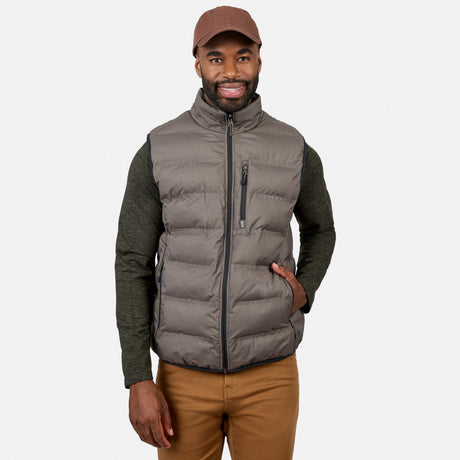 A man wearing the Free Country Men’s Haven Stitchless Reversible Vest in gray, along with a brown cap, green long-sleeve shirt, and tan pants, stands smiling with one hand in his pocket against a plain white background.