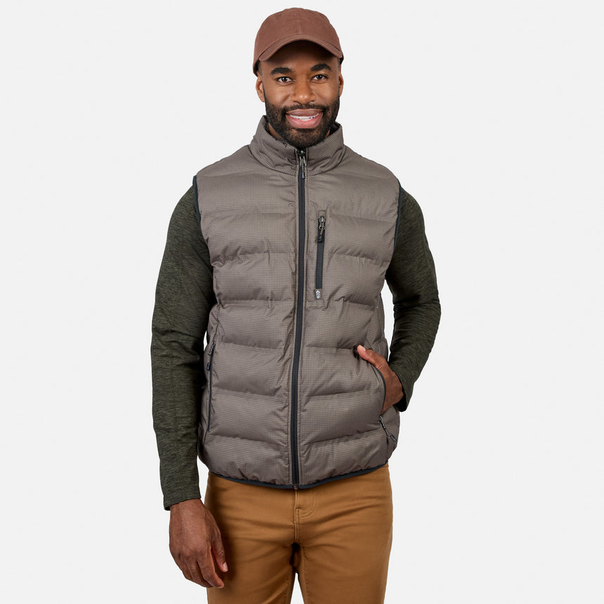 A man wearing the Free Country Men’s Haven Stitchless Reversible Vest in gray, along with a brown cap, green long-sleeve shirt, and tan pants, stands smiling with one hand in his pocket against a plain white background.