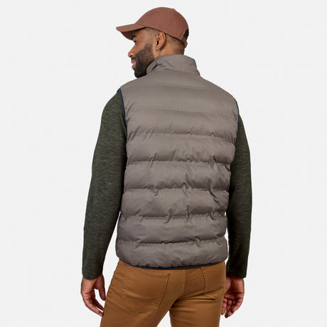A man is standing and facing away from the camera against a plain white background, wearing a brown cap, green long-sleeve shirt, brown pants, and the Free Country Men’s Haven Stitchless Reversible Vest in gray, which provides lightweight warmth.